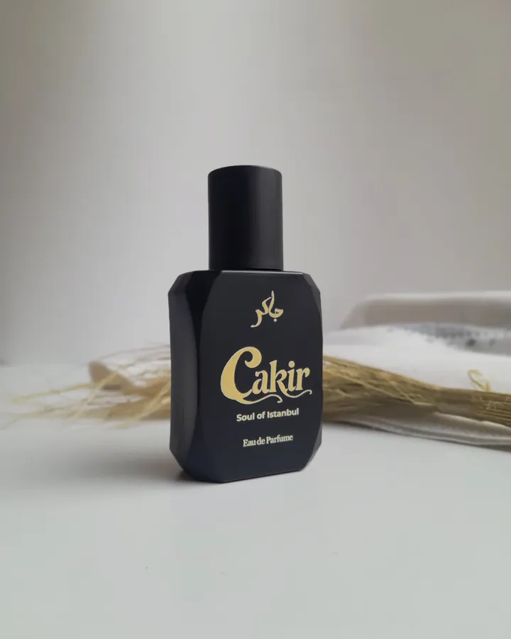 Cakir Parfume V5.2 — black bottle with dried lemongrass and warm cream pillow, soft natural light